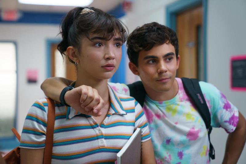 In a still from the movie Crush, Rowan Blanchard as Paige is in a stripped t-shirt and has her hair in a scrunch. She looks bewildered while her best friend, Tyler, who is wearing a tie dye shirt, has his arm around her shoulder.