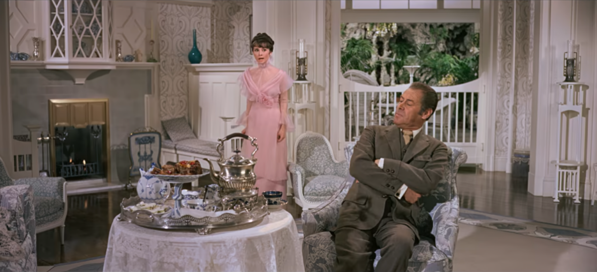 In the movie My Fair Lady, Audrey Hepburn us wearing a pink dress and standing. Rex Harrison is sitting at a table set for tea service and leaning away from it while folding his arms.