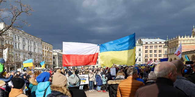 A crowd gathers outside on a cloudy day. In the middle are large flags, one of Poland and the other of Ukraine.
