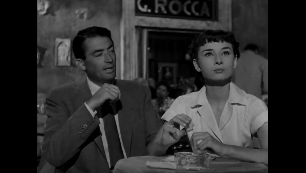 In the black-and-white film Roman Holiday, Gregory Peck wears a suit while looking at Audrey Hepburn, who is wearing a white collared dress.