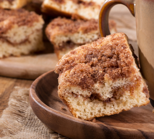 A square slice of cinnamon coffee cake is on a wooden tray next to a brown mug. In the background, there is a pile of other coffee cake slices.