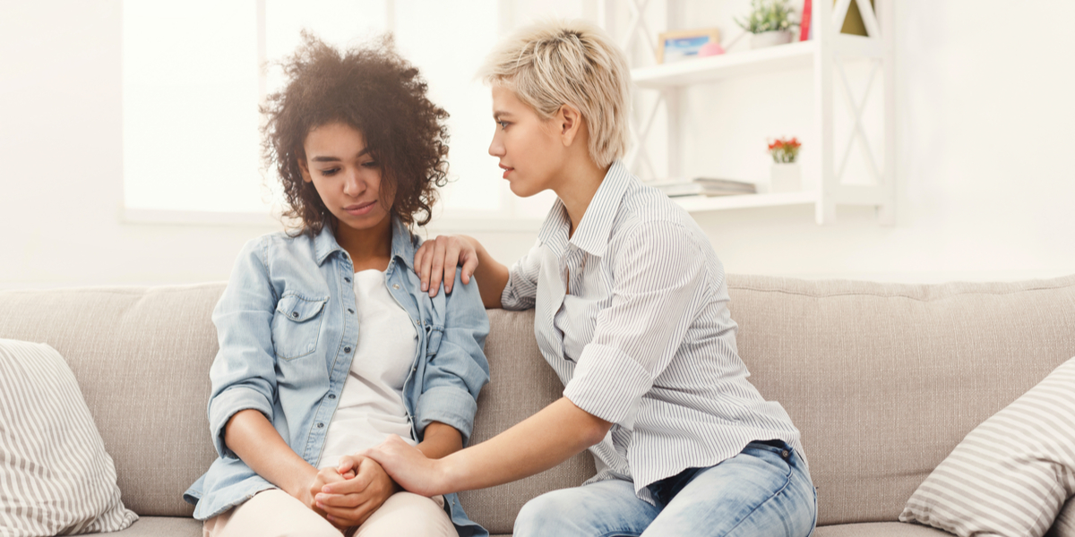 two women sit on a couch talking to each other, looking serious and maybe a little sad