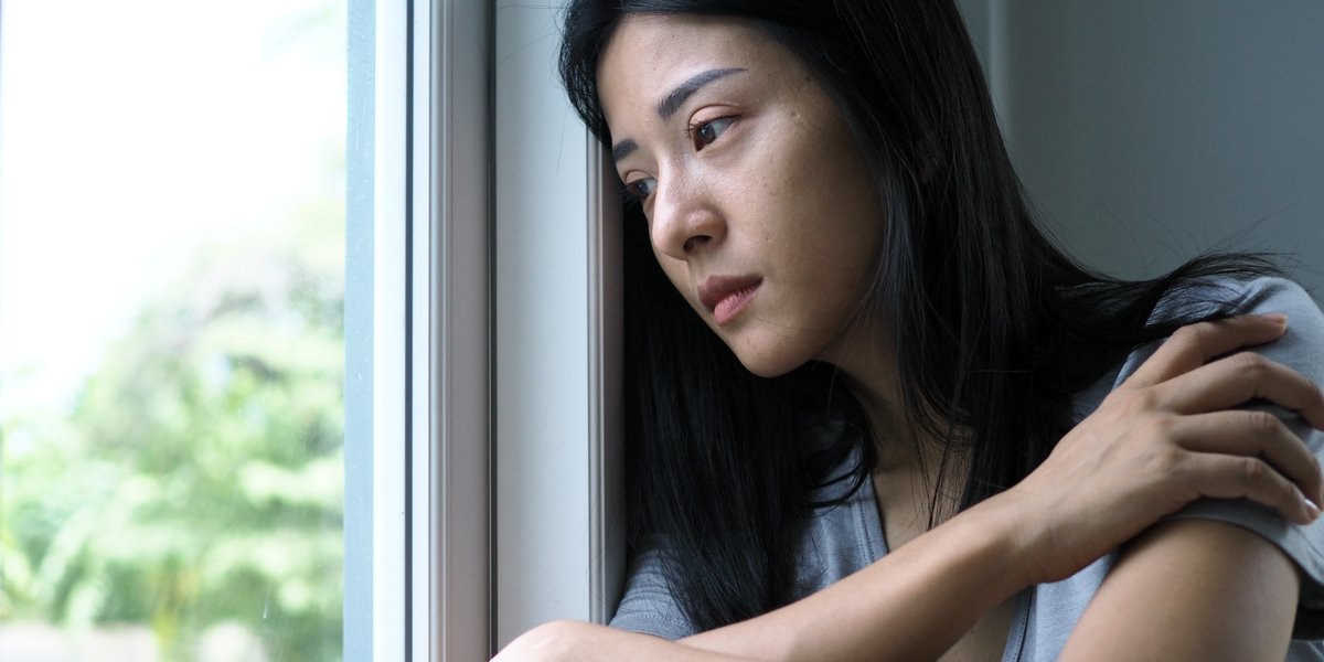 An East Asian woman with shoulder-length black hair and brown hairs stares out a window with her arms wrapped around her chest. Trees are visible through the window.