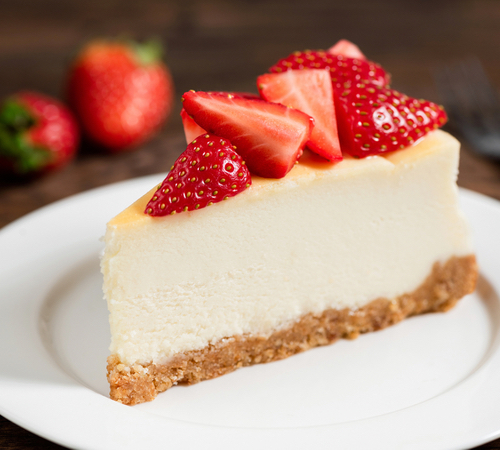 A slice of cheesecake topped with strawberries is on a white plate. Two whole strawberries are visible in the background.