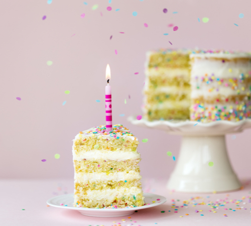 Against a pink background, a slice of layered confetti cake with white frosting sits on a small, white plate. A pink, lit candle is in the slice. In the background, the rest of the cake is on a white cake stand. Multi-colored confetti falls in front of the image.
