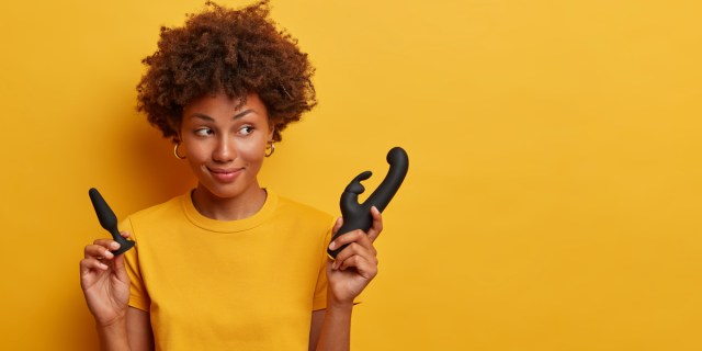 A Black woman with a reddish-brown afro wears gold hoop earrings and an orange T-shirt. She stands in front of an orange background and holds a black butt plug in one hand and a black rabbit vibrator in the other.