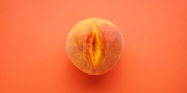 Against an orange background, there is a peach with a slit cut down its center, causing the fruit to resemble a vulva.