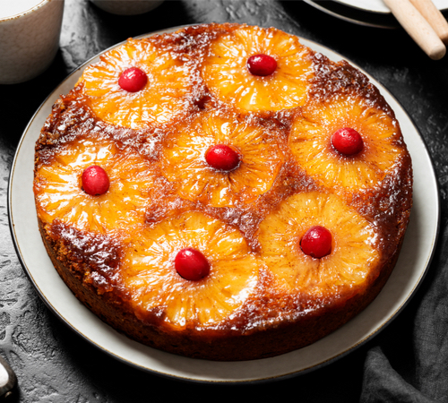 A pineapple upside-down cake topped with pineapple rings and cherries is on a white plate.
