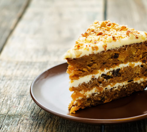 A slice of carrot cake with white frosting sits on a brown plate on a wood table.