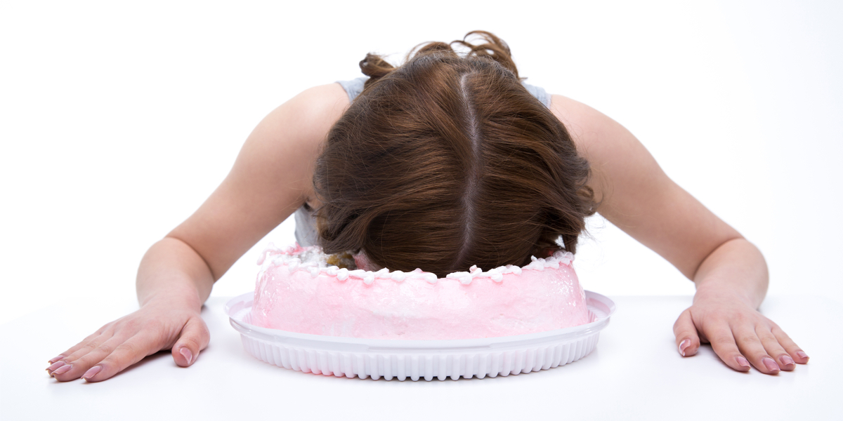 Against a white background, a white woman with shoulder-length brown hair leans over a white table and presses her face into a pink cake.