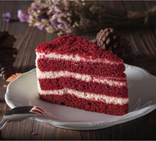 A slice of dark red cake with layers of white frosting is on a white plate on a wood table. A spoon rests on the plate. Pinecones are scattered in the background.