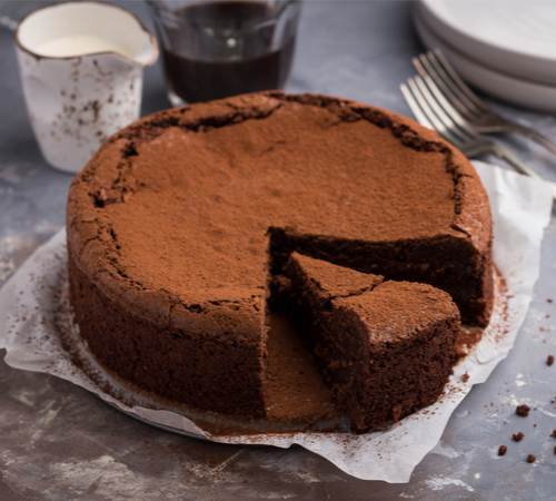 A flourless chocolate cake with a slice cut out of it is on a sheet of wax paper. In the background, a white pitcher of cream, a glass mug of coffee and two forks are visible.