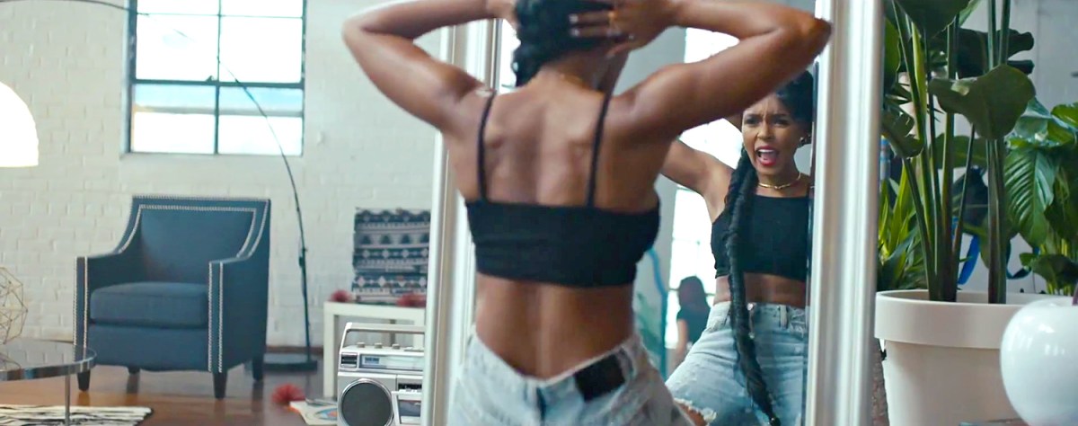 Janelle Monáe in her music video "Yoga" sits in front of a mirror in a black crop top and jeans while flexing her back/shoulder muscles.