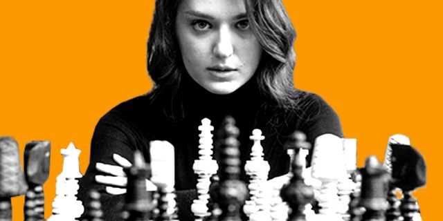 A white person with mid-length wavy hair sits, with focused eyes and parted lips, behind a chess board