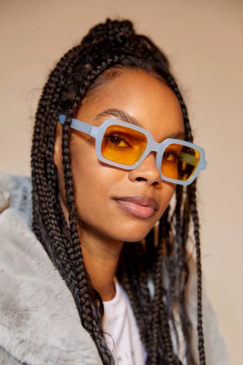 A black woman with long braids in a grey sweater coat has on light blue square sunglasses with orange lenses.