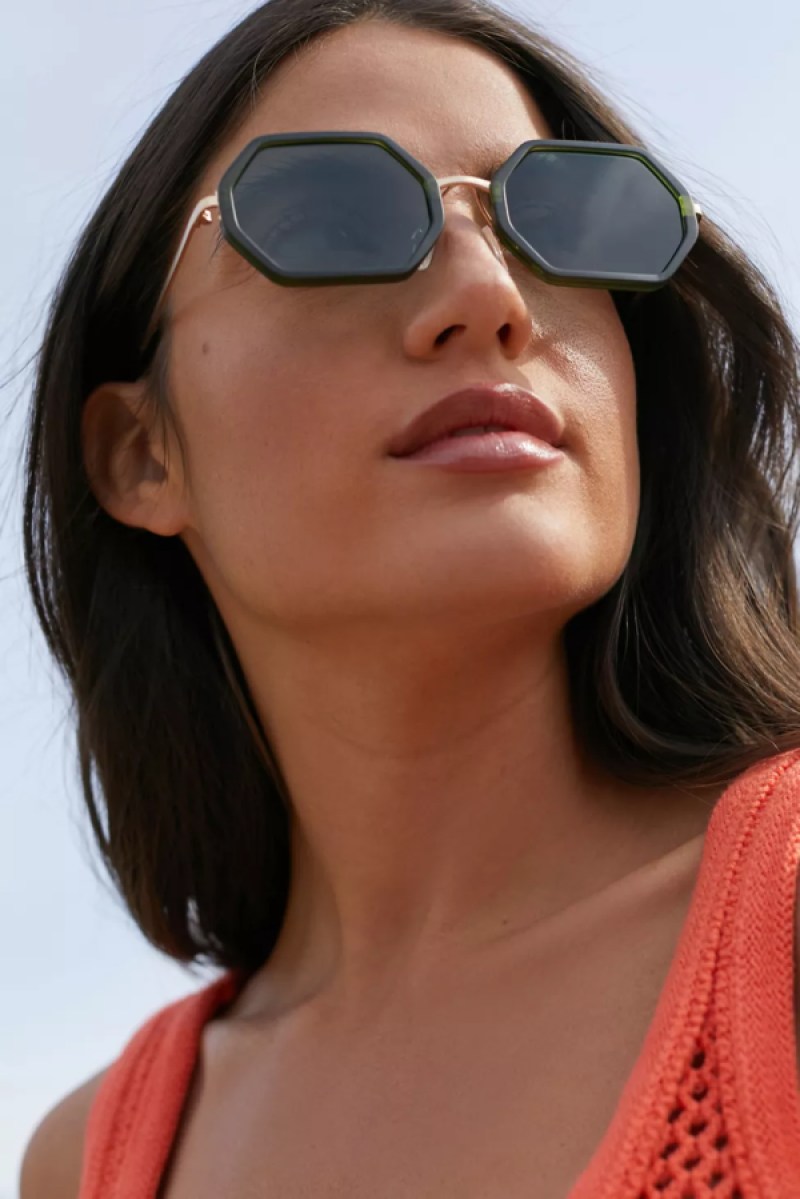 A woman with brown skin, long dark hair, and an orange tank top has on dark green hexagon shaped sunglasses with thin gold frames. 