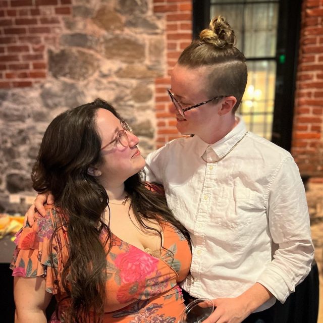 Vanessa and PB pose in formalware at some kind of event. Vanessa is wearing a bright floral dress, with her long brown hair down. Vanessa is a white woman with glasses. PB is taller than Vanessa, and is wearing a white button up shirt with a chain necklace and dark glasses. She is a white woman with her long blonde hair in a bun on the top of her head with her sides shaved.