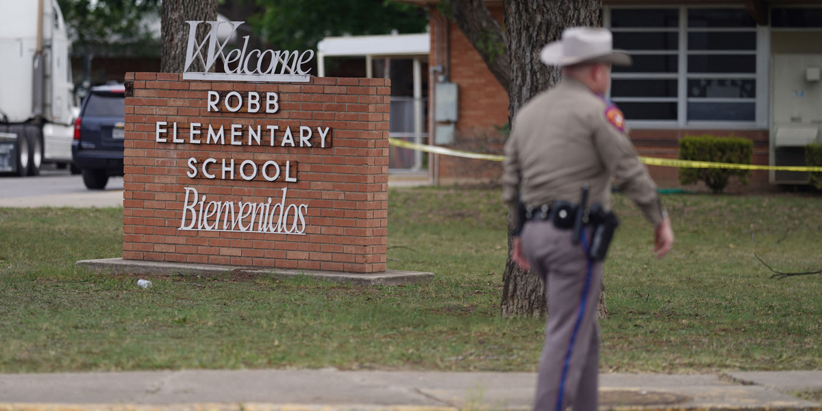A school sign for Robb Elementary Schools is brick and says Bienvenidos in white cursive. In front of the sign is a Texas sheriff with his back to the camera, out of focus.
