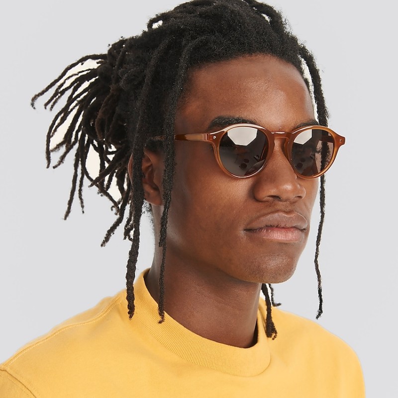 A black masc person with dreadlocks in a messy top bun and a yellow shirt wears round glasses with a keyhole on the nose bridge, the glasses have dark lenses and a tan brown frame.