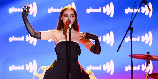 Dove Cameron sings on stage at the GLAAD Awards in front of a blue background, she's in a sleeveless black dress with long opera gloves up to her elbow.