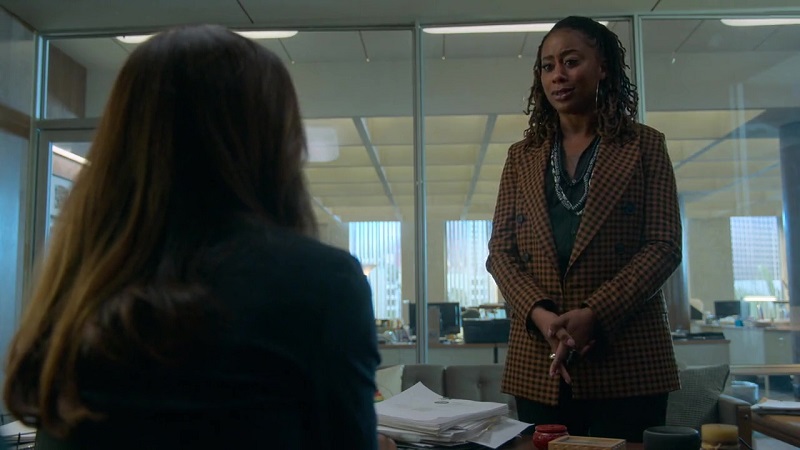 Malika steps in to Lucia's office to apologize for her actions and try to save her job. She's standing in front of Lucia's desk, wearing a gingham blazer with green blouse.