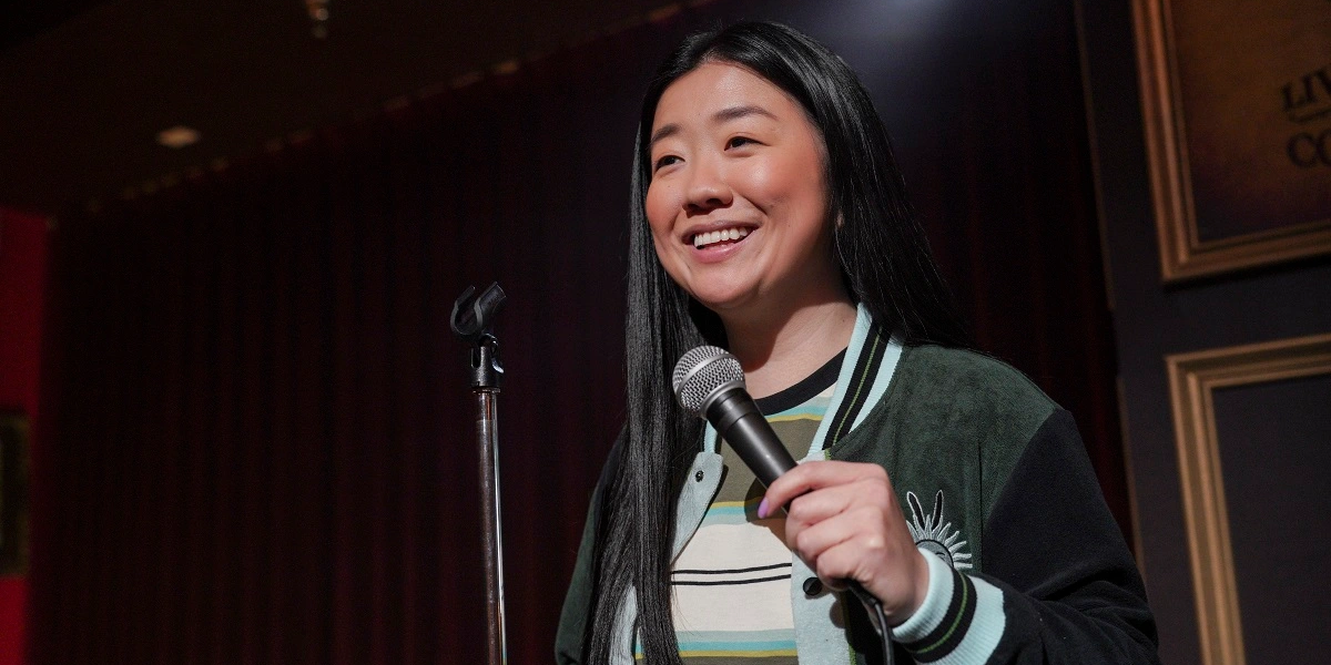 Alice gets back on stage after her harrowing encounter on tour. She's holding a mic in her left hand and wearing a striped shirt and varsity jacket.