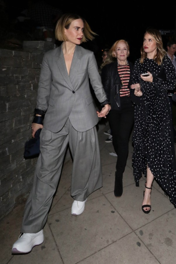 Sarah Paulson wears a gray suit and white sneakers while walking ahead of Holland Taylor, whose hand she is holding. Holland wears a striped shirt, black blazer, and black pants. Leslie Grossman is also walking with them, wearing a long black dress with white polka dots.