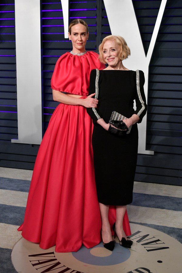 Sarah Paulson and Holland Taylor stand together. Sarah is wearing a long red dress, and Holland is wearing a black dress with a square neck. Sarah is holding Holland's arm.