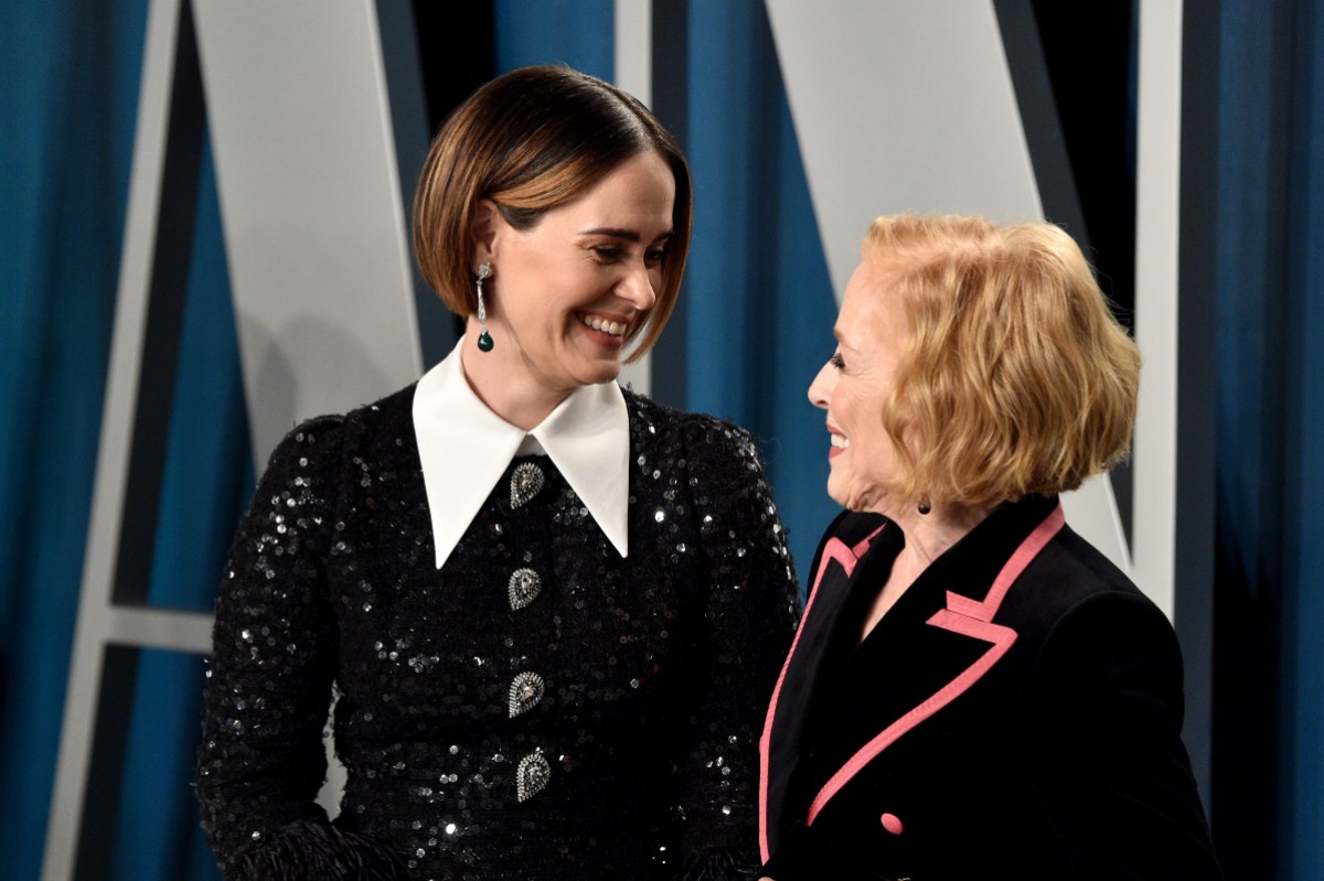 Sarah Paulson and Holland Taylor are gazing into each other's eyes while smiling. Sarah wears a sparkly black dress with a white collar. Holland wears a black suit with pink detailing.