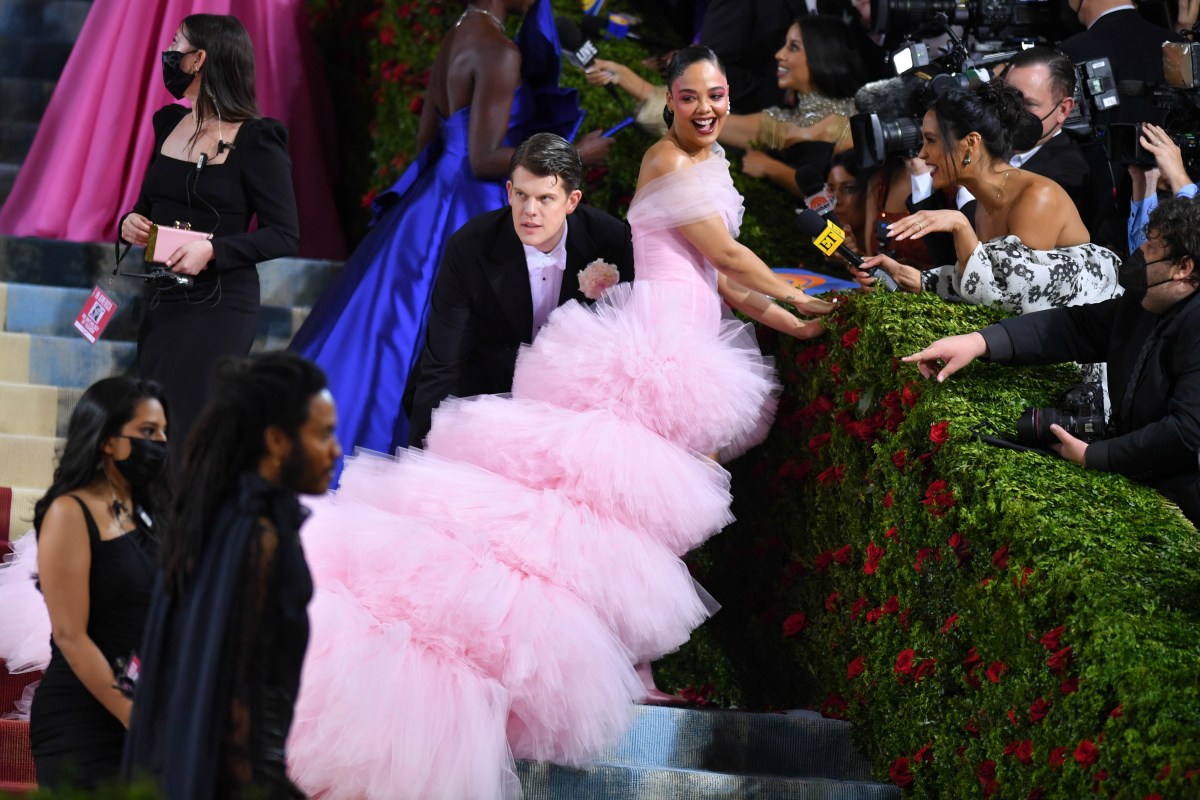 Tessa Thompson climbs the stairs on the red carpet at the Met Ball, she's in a 5 tier fluffy pink gown that looks like a five tier cake (it does!), she is smiling.