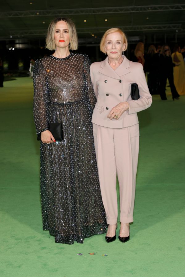 Sarah Paulson and Holland Taylor stand together. Sarah is wearing a long sparkly black dress and holding a clutch. Holland is wearing a beige double breasted jacket suit.