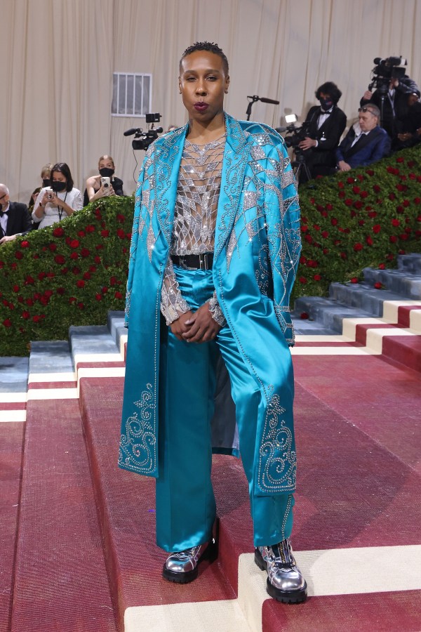 Lena Waithe in a blue silk suit styled like Elvis with bedazzled jewels. She has dark brown lipstick and is posing with one leg up and her hands between her legs.
