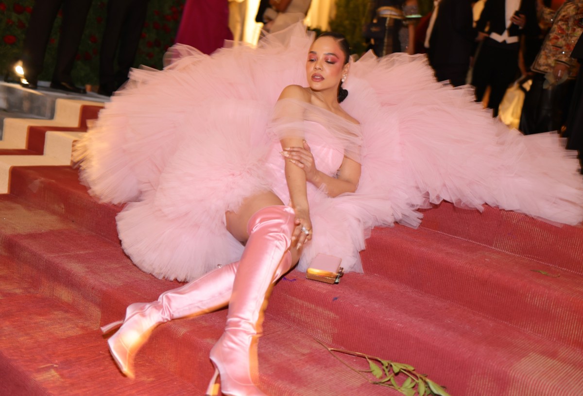 Tessa Thompson is sitting on a couch with her feet propped up underneath her fluffy pink gown, showing off thigh high satin pink boots