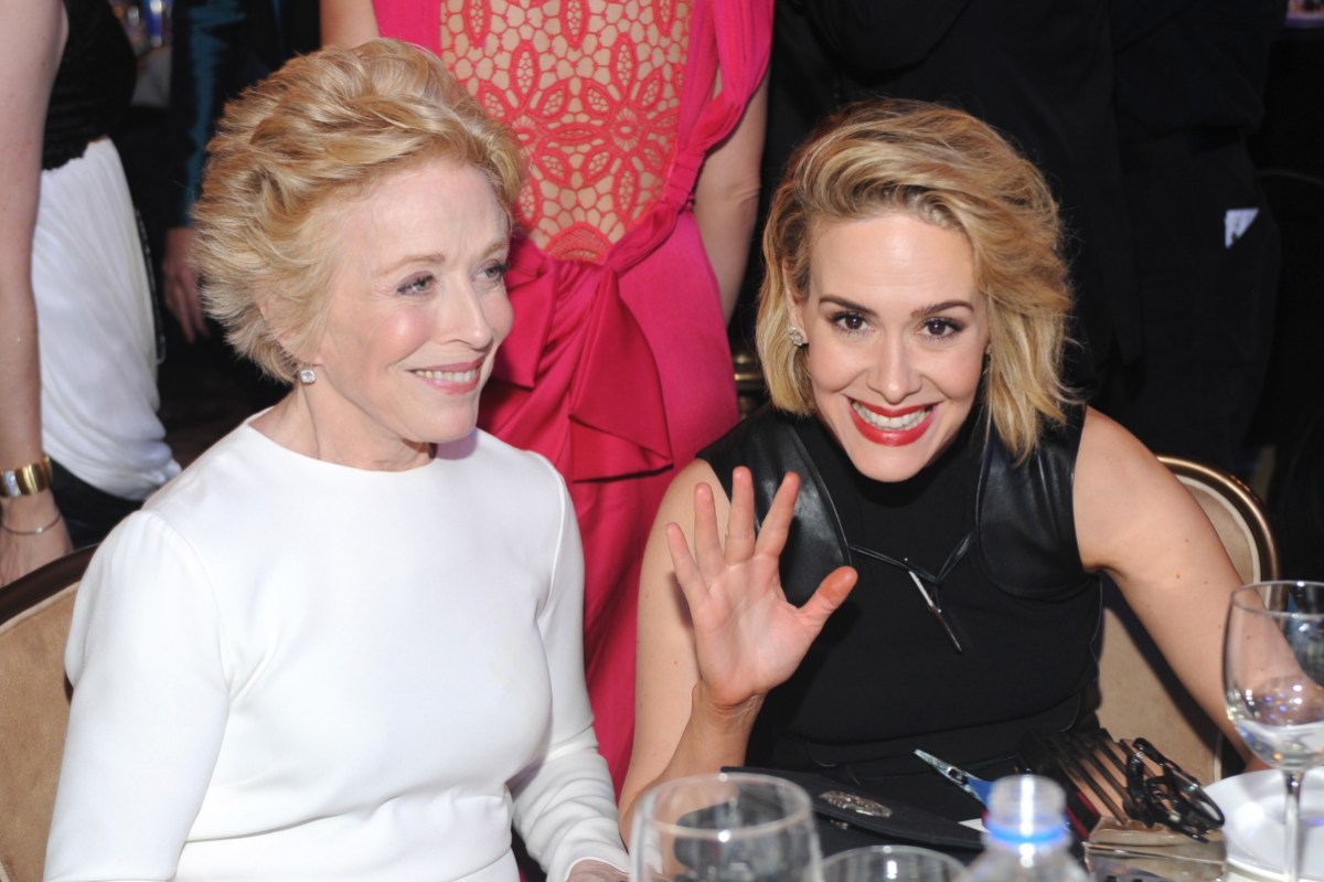 Holland Taylor and Sarah Paulson are sitting at a table. Holland is wearing a white dress with long sleeves. Sarah is wearing a black sleeveless dress and smiling while waving at the camera.