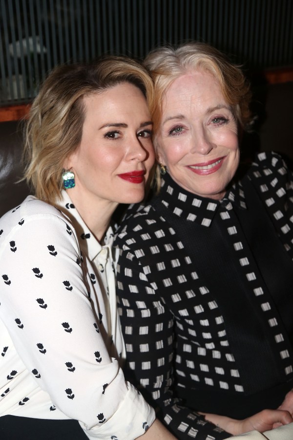 Sarah Paulson and Holland Taylor are snuggling. Sarah is wearing a white buttondown blouse with a black floral print on it. Holland is wearing a checkered suit set. They are both smiling at the camera.