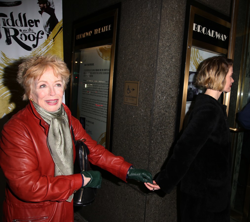 Sarah Paulson and Holland Taylor are holding hands while walking. Sarah is walking ahead of Holland and wearing all black while looking away. Holland is wearing a red leather coat and beige scarf and walking behind Sarah. A sign for Fiddler on the Roof is in the background.