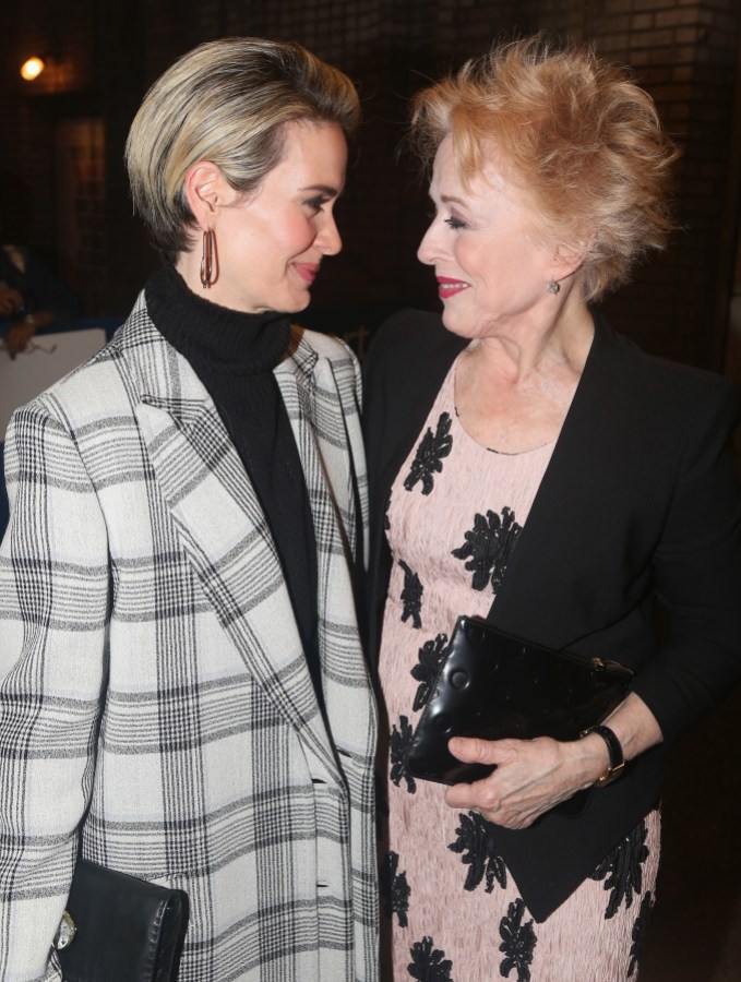 Sarah Paulson and Holland Taylor stand close to each other while looking in each other's eyes. Sarah is wearing an off white and black checked wool coat and a black turtleneck. Holland is wearing a pale pink dress with black detailing and a black cardigan.