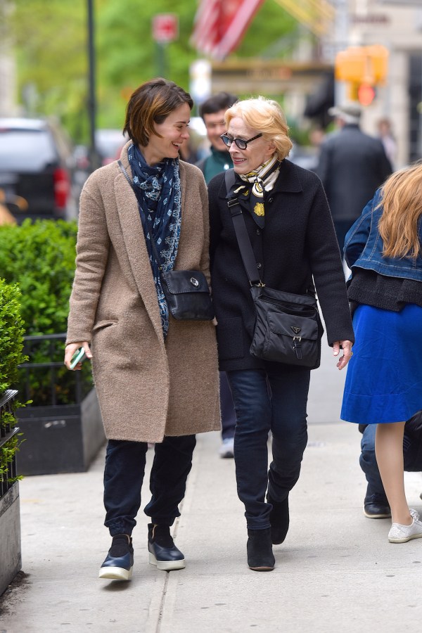 Sarah Paulson and Holland Taylor are walking on a sidewalk. Sarah is wearing a long light brown teddy bear coat, black slouchy pants, and a big blue scarf. Holland is wearing a black jacket, black pants, and a patterned silk scarf. They both have black cross-shoulder bags. They're talking to each other and smiling.