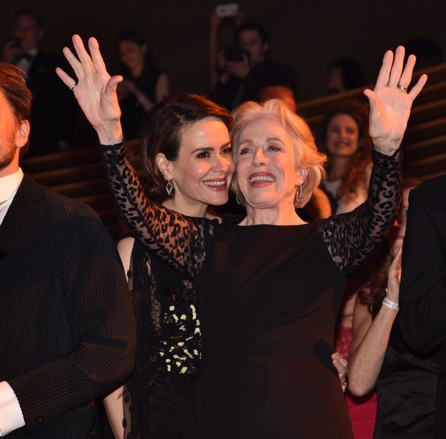 Holland Taylor is wearing a black dress and holding her hands up in the air. Sarah Paulson is standing behind her, smiling, also wearing a black dress.