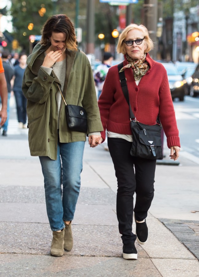 Sarah Paulson and Holland Taylor are holding hands while walking. Sarah is biting her hand and looking down while wearing a large olive green coat, jeans, and a cross-shoulder bag. Holland Taylor is looking at the camera, wearing glasses, a red cardigan, black pants, and black boots.