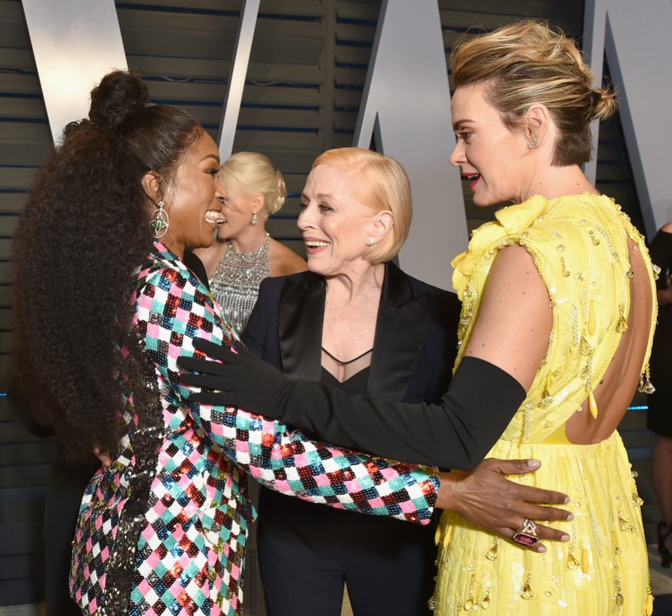 Angela Bassett, Holland Taylor, and Sarah Paulson are at a red carpet event. Angela wears a colorful, shiny suit with a checked pattern and a large cocktail ring. Sarah wears black elbow-length gloves and a yellow evening dress. Holland wears a black suit. Angela's hand is on Sarah's butt.