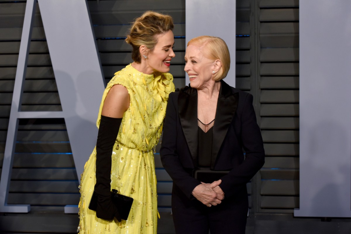 Sarah Paulson stands with Holland Taylor, looking at her and smiling. Sarah wears black elbow-length gloves and a yellow evening dress. Holland wears a black suit.