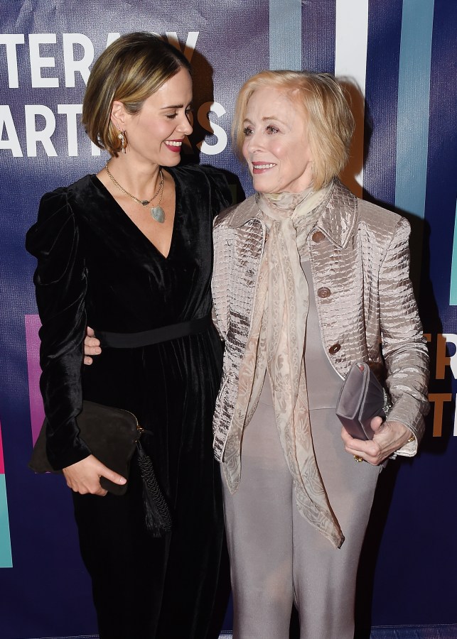 Sarah Paulson and Holland Taylor stand with their arms around each other. Sarah is giving Holland googly eyes. Sarah wears a black dress with a v-neckline, and Holland wears a taupe suit.