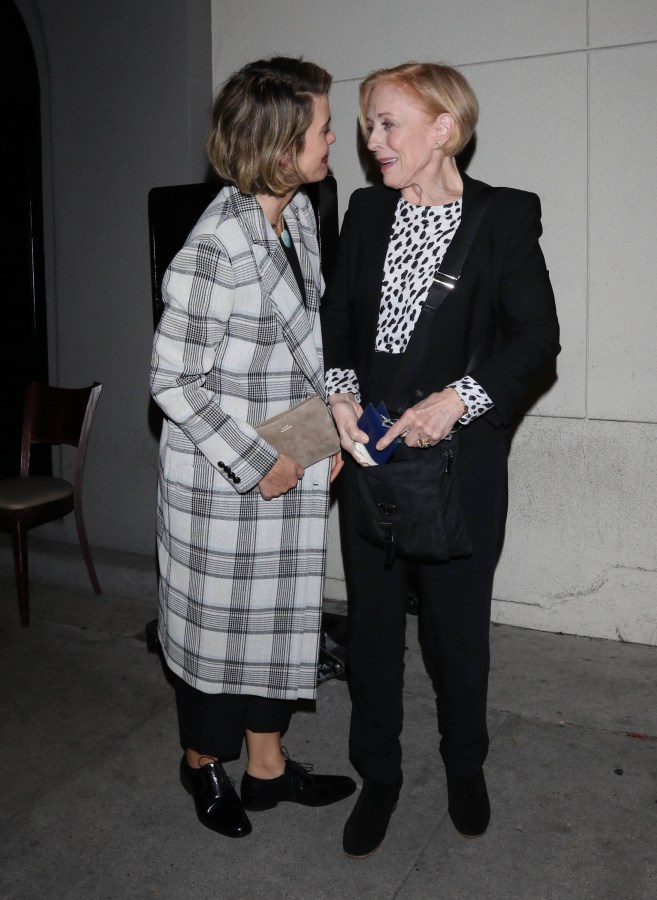 Sarah Paulson and Holland Taylor stand while looking in each other's eyes. Sarah is wearing a long plaid wool coat, and Holland is wearing a black suit with a black and white spotted blouse.
