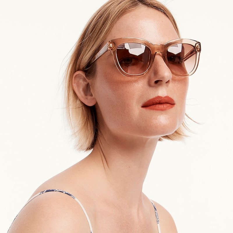 A white femme person with rust colored lipstick and a blonde bob wears oversized square sunglasses with both lenses and frames in a monotone tan color.