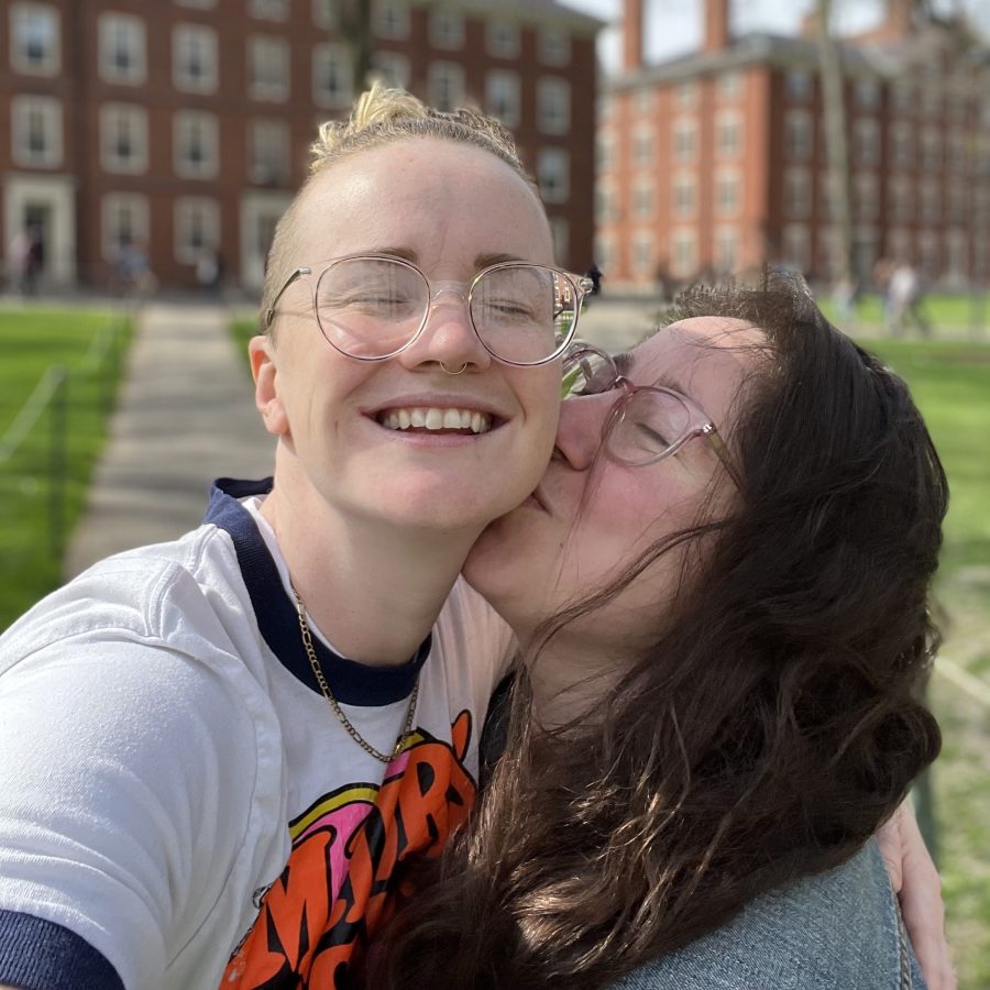 PB grins widely while Vanessa kisses her on the cheek. PB is a tall white woman with blonde hair that is long on top and shaved on the sides who wears clear glasses and is here wearing a tee shirt. Vanessa is shorter than PB, and is a white woman with long brown hair and pink tinted glasses. 