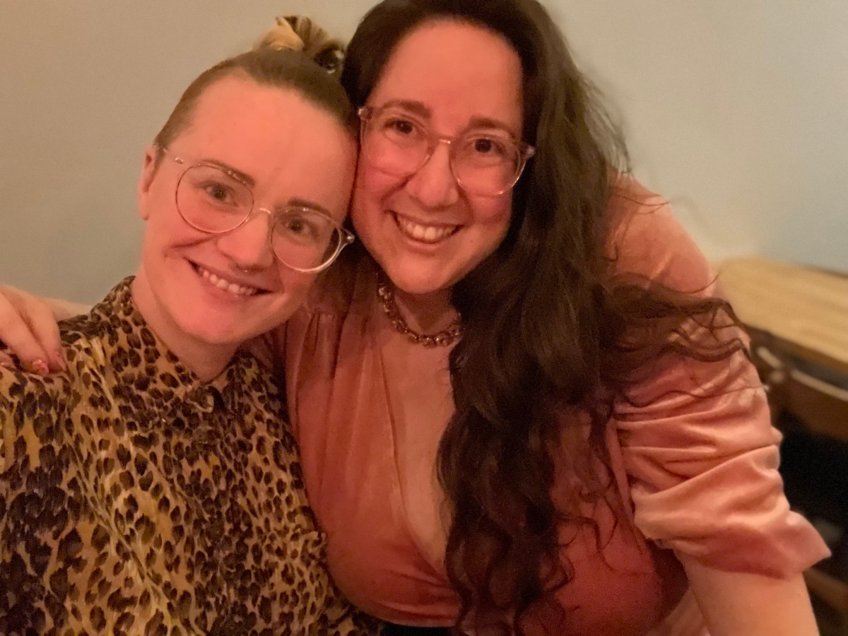 PB and Vanessa pose for a photo while smiling. PB is wearing glasses and a cheetah print button up. Vanessa is wearing a pink velvet dress and necklace. PB is white and has blonde hair that is long on top and up in a bun with shaved sides. Vanessa is also white with long brown hair she is wearing down.