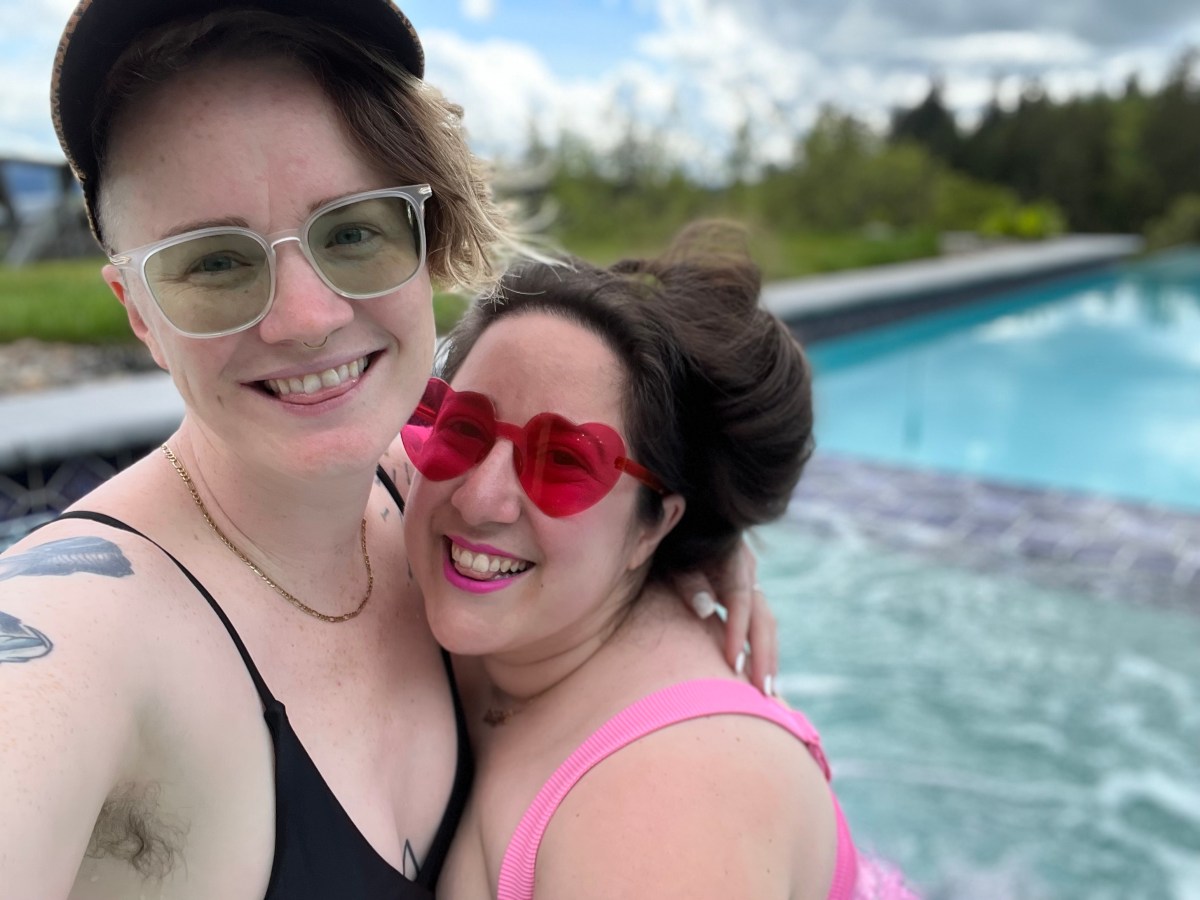 Vanessa and PB pose in front of a pool in bathing suits, wearing sunglasses. Vanessa's sunglasses are red and heart-shaped. PB's are clear with greenish lenses. PB is white and has blonde hair that is long on top and under a hat with a swoop of bangs visible and with shaved sides. Vanessa is also white with long brown hair she is wearing up.