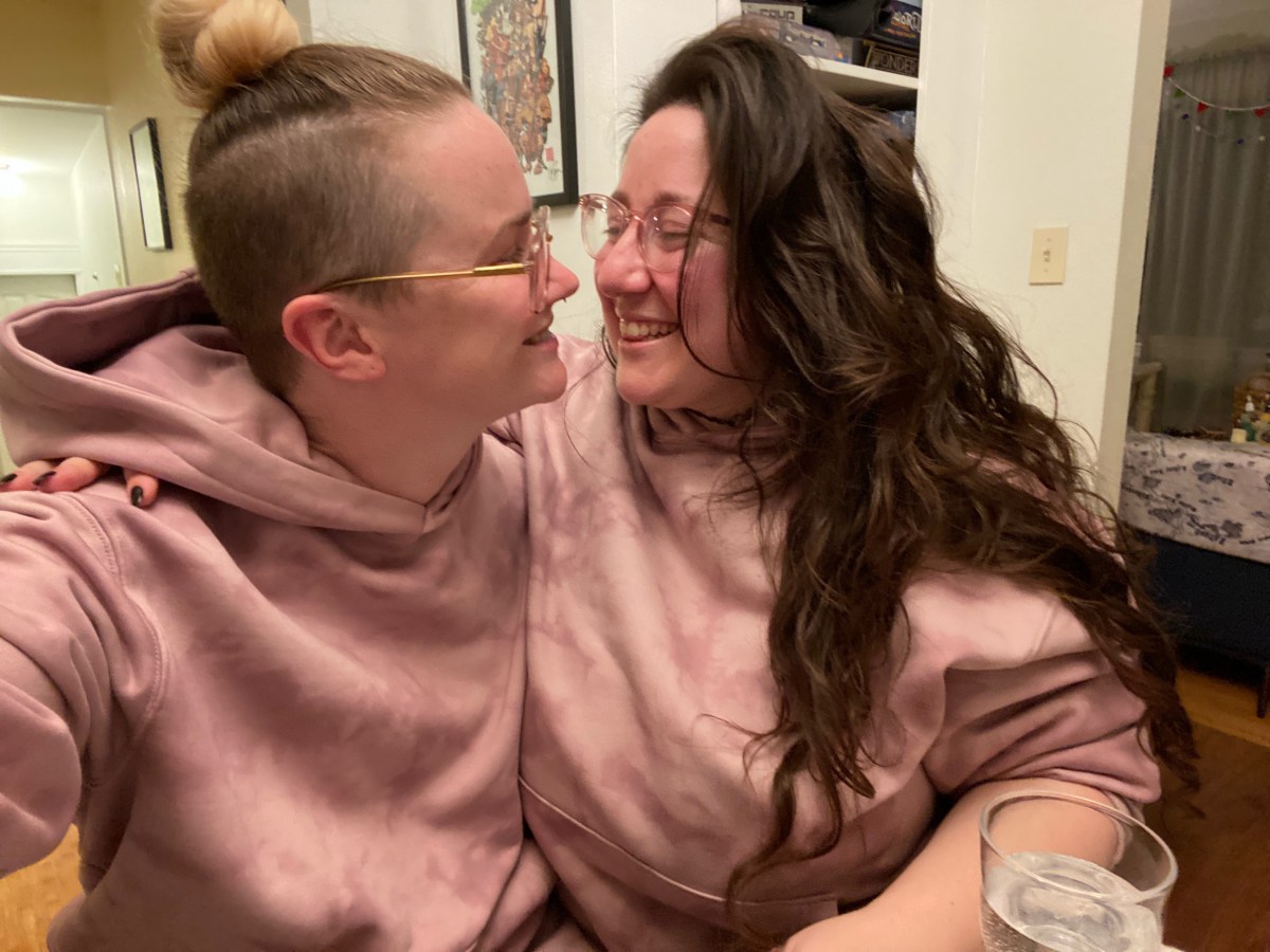 PB and Vanessa sit in matching purple sweatsuits with their arms around each other, gazing lovingly into each others eyes. They are also both wearing glasses. PB is white and has blonde hair that is long on top and up in a bun with shaved sides. Vanessa is also white with long brown hair she is wearing down.