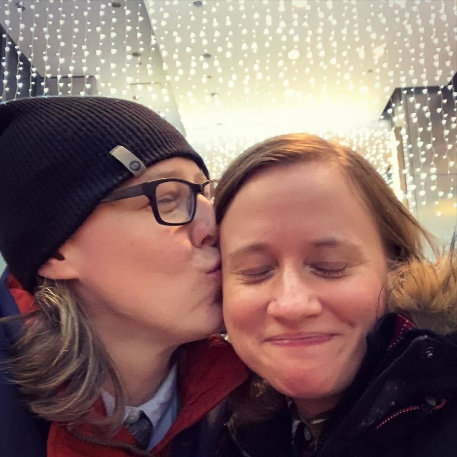 Heather and Stacy are pictured in this photo. In the photo, Heather, a soft butch white woman wearing black rimmed glasses and a beanie, kisses Stacy, a white woman with light brown hair, on the cheek. They are both smiling and both look happy. You can see string lights glowing behind them..